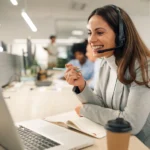 Smiling caucasian business woman with headset working on a laptop in a call center on the line Smiling caucasian business woman with headset working on a laptop in a call center on the line. Happy female working in a busy customer service department and taking notes while talking with a client